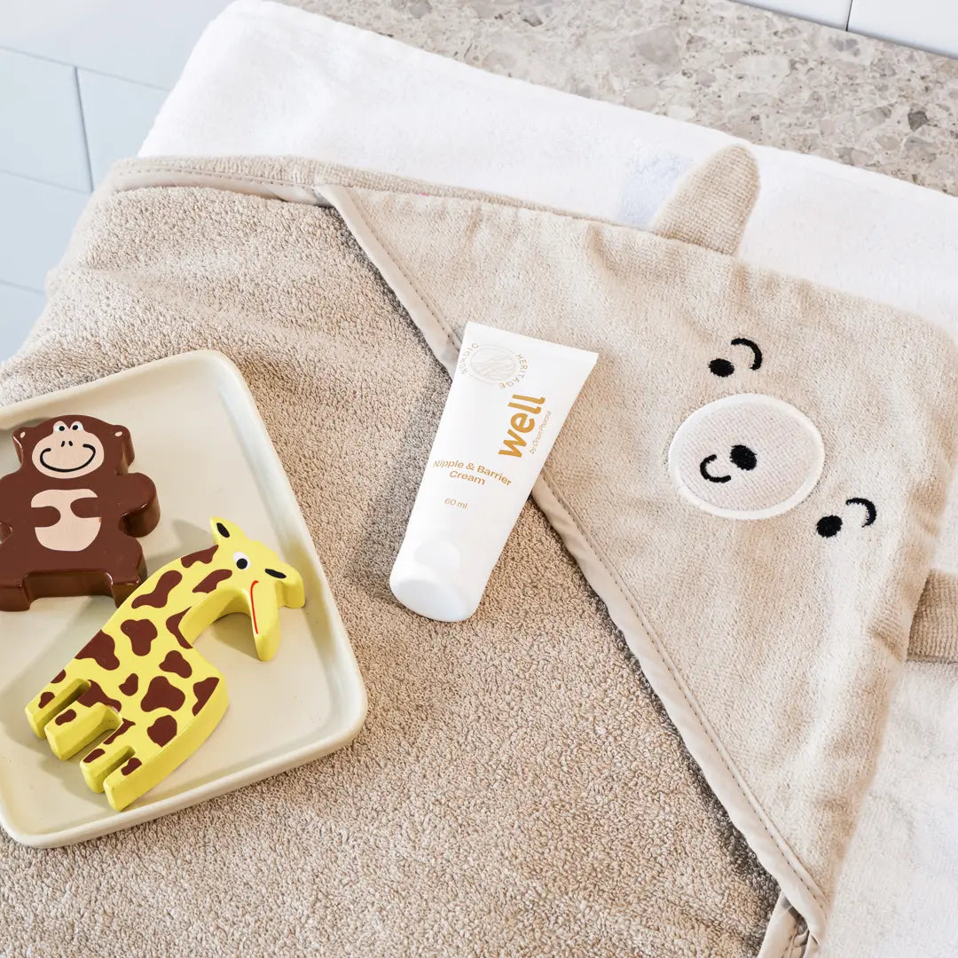 Beige towel with cartoon face, white tube labeled "Well by Orion Pharma", and animal-shaped toys on a light wooden surface.