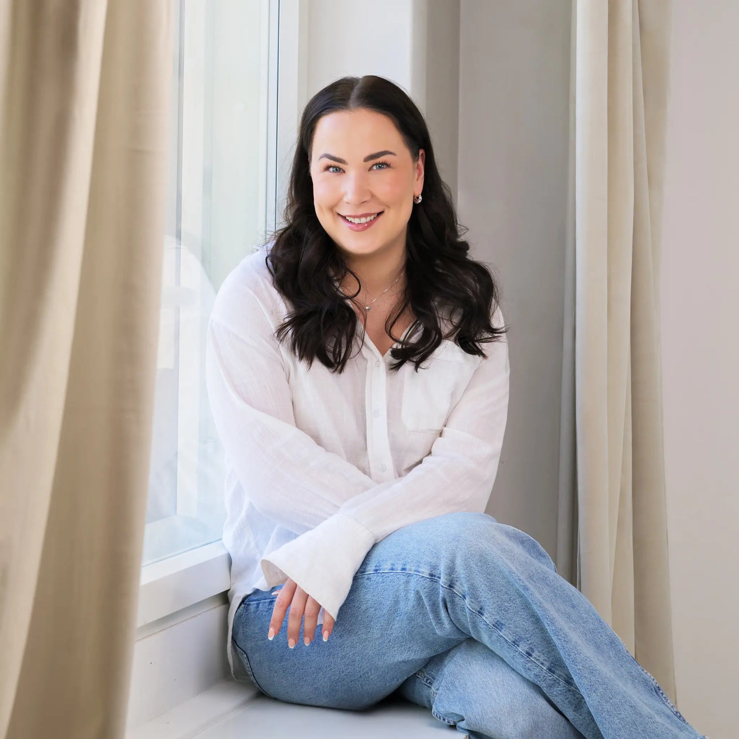 Woman sitting by a window wearing a white blouse and blue jeans.