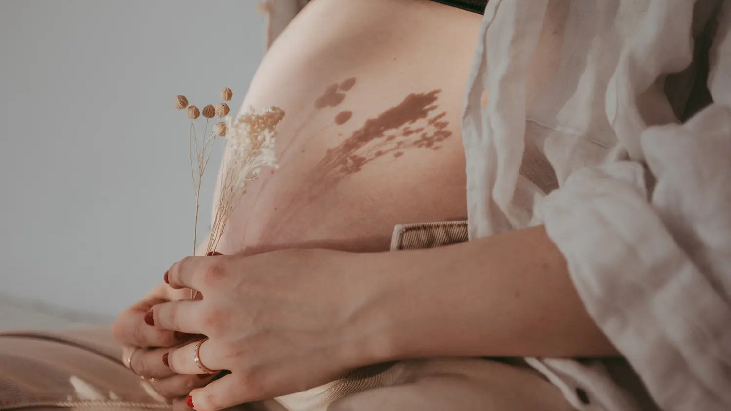 Close-up of a pregnant belly with hands gently holding it, surrounded by dried plants.