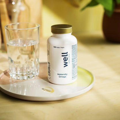 Bottle of 'well' Maternity Omega supplement on a white plate with a glass of water, on a wooden surface.