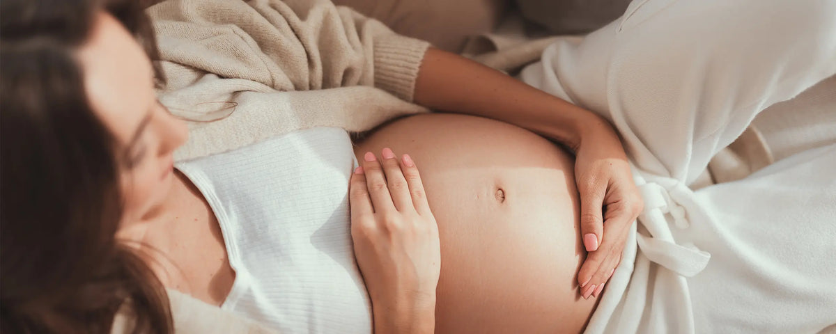 Närbild av en gravid kvinna med en hand på magen, klädd i vitt.