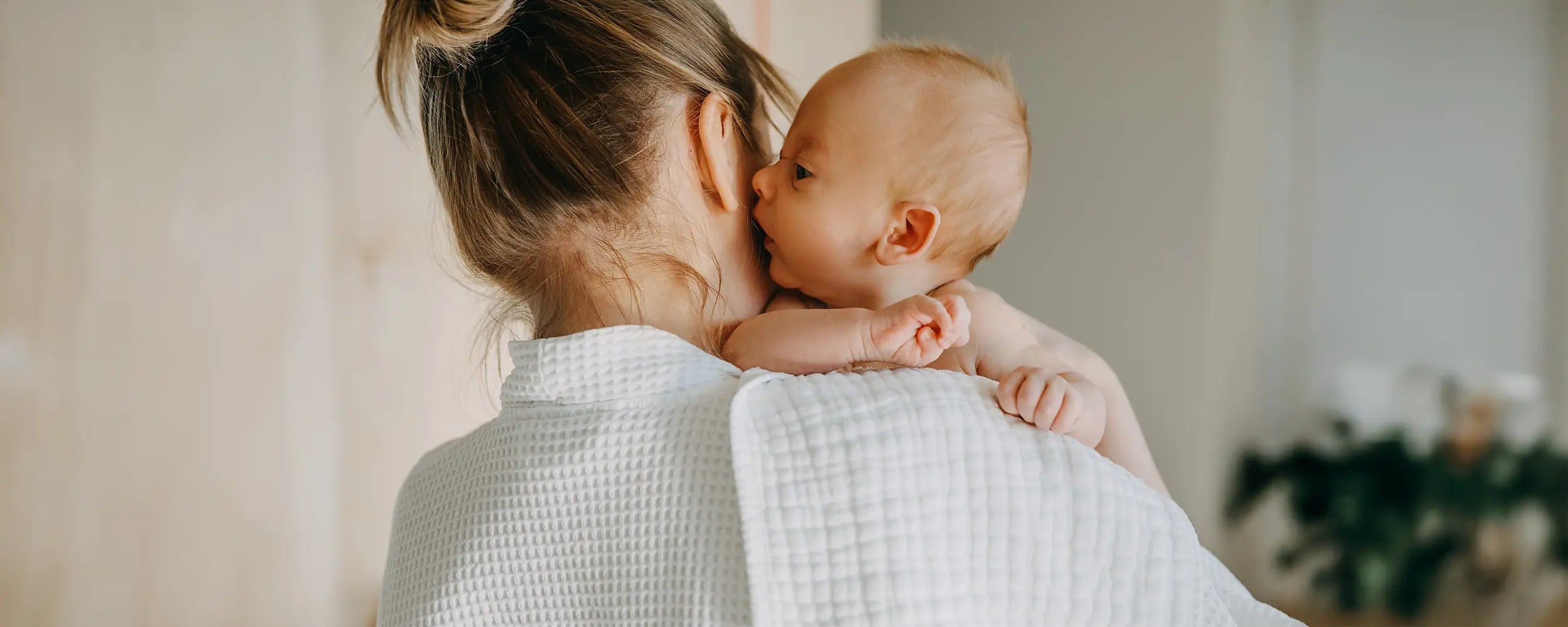 Woman holding a baby wrapped in a white blanket indoors.